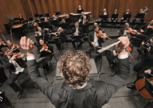 Downward shot of the top of an orchestra conductor's head of brown hair, while he conducts several musicians dressed in black who are seated in front of him.